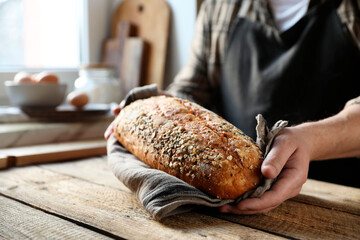 Man holding loaf of fresh bread at wooden table indoors, closeup