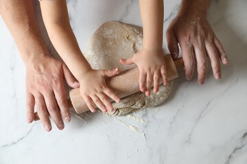 Father and child rolling raw dough at white table, top view