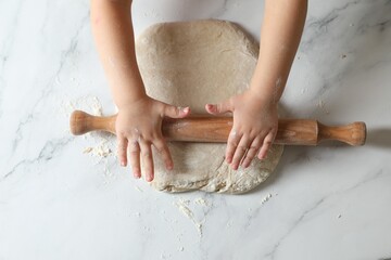 Little child rolling raw dough at white table, top view