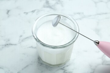 Mini mixer (milk frother) and whipped milk in glass on white marble table, closeup