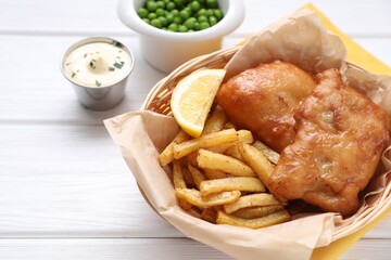 Tasty fish, chips, sauce and lemon in wicker bowl on white wooden table, closeup. Space for text