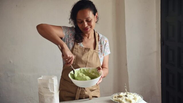 Pretty woman housewife baking at home. Female chef in beige apron, kneading dough for making homemade ravioli, preparing delicious dumplings with mashed potatoes for family dinner in the rural kitchen