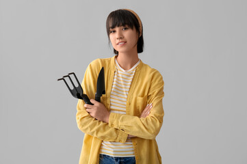 Beautiful young happy female gardener with gardening rake and shovel on grey background