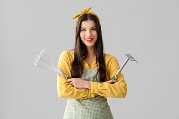 Beautiful young happy woman with gardening rakes on grey background