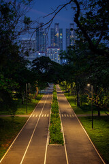 Lit skyscrapers behind a pathway or running track and lush trees at the Benjakitti Forest Park in Bangkok, Thailand at dusk.