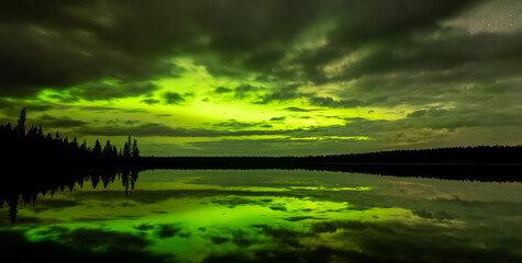 Bright green aurora fill the sky behind clouds above a calm northern lake. The quiet water has a...