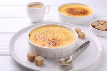 Delicious creme brulee in bowls, vanilla pods, sugar cubes and coffee on white wooden table, closeup