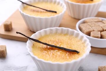 Delicious creme brulee in bowls, vanilla pods and sugar cubes on white marble table, closeup