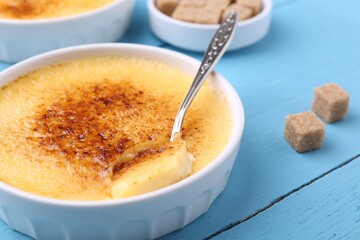 Delicious creme brulee in bowl, sugar cubes and spoon on light blue wooden table, closeup