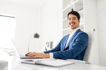 An affable Hispanic businessman smiles, looks at the camera while using a laptop in a well-lit,...