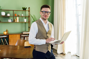 A professional man in a vest stands confidently in his well-lit office, balancing a laptop,...