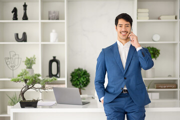 Young businessman talking on the phone while holding hand in pocket standing in bright office, showcasing a dynamic work environment where multitasking is the norm