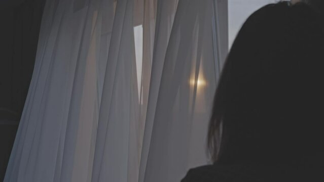 Woman opening curtains to let in bright morning sunlight in bedroom. Person pulls back light curtains and looks at sunrise in empty city streets
