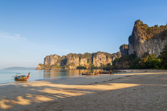 Scenic view of few long-tail boats, Railay West beach and high limestone cliffs in Railay, Krabi, Thailand, on a sunny morning.