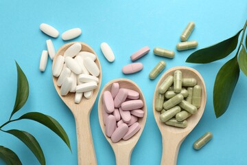 Different vitamin pills in spoons and green leaves on light blue background, flat lay