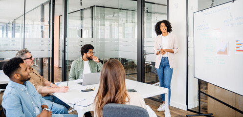 A charismatic professional woman stands presenting at a whiteboard, fostering an interactive brainstorming session in a modern office. The diverse team, attentively listening and taking notes - obrazy, fototapety, plakaty