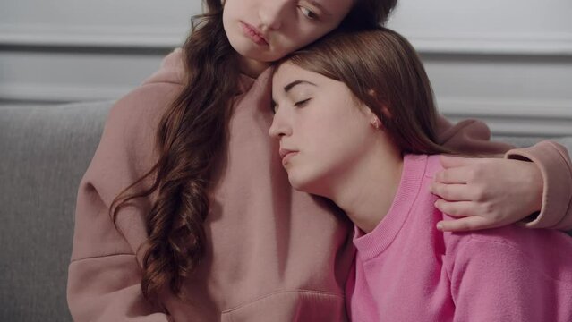 Two cute girls teenagers or students in sad emotions sit on the sofa, hug and comfort each other. A young lady strokes friend or sister on the shoulder, kisses forehead, supports in a difficult