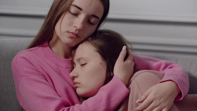 Two cute girls teenagers or students in sad emotions sit on the sofa, talk, hug and comfort each other. A young lady strokes friend or sister on the head and shoulder, supports in a difficult