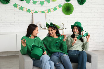 Beautiful young women with decorative glasses in shape of clover on sofa at home. St. Patrick's Day celebration