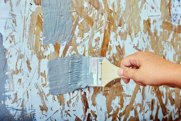 House painter applies brush strokes with gray paint to chipboard wall.