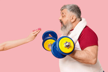 Overweight mature man with dumbbells and hand holding donut on pink background. Weight loss concept