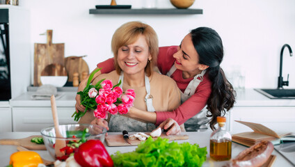 Happy daughter congratulates mom and gives her tulips at home kitchen while she cooking. Birthday, Mothers day, women's day, retired, family, relation, motherhood. - 773485933