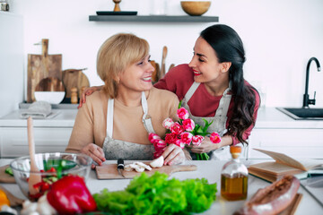 Happy daughter congratulates mom and gives her tulips at home kitchen while she cooking. Birthday, Mothers day, women's day, retired, family, relation, motherhood.