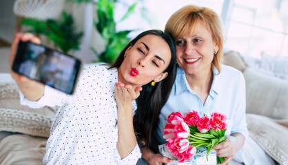 Happy mother and daughter making photo on smart phone with flowers at home on the couch. Birthday, Mothers day, women's day, retired, family, relation, motherhood. - 773486124