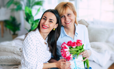 Excited mother and daughter with present and flowers at home on the couch. Birthday, Mothers day, women's day, retired, family, relation, motherhood. - 773486131