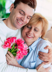 Close up vertical portrait of young son hugging his smiling mother at home with bouquet of tulips. Birthday, Mother's day, women day, retired. - 773486173