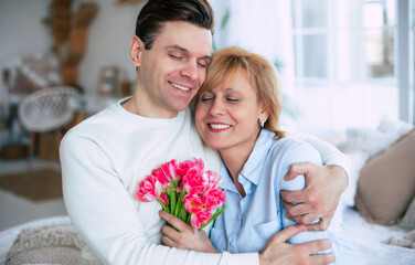 Surprise in Mother's day. Handsome son embracing his happy mother at home with bouquet of tulips - 773486177