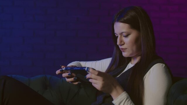 Young woman sitting on couch and playing portable console with neon light background. Playing games alone at home. Female sitting on sofa, concentrated on match in video game. Concept of portable