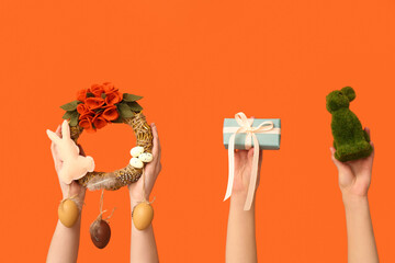 Female hands with Easter festive wreath, bunny and gift box on orange background