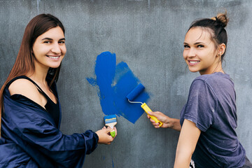 Painting work, smiling young women paint gray wall with blue paint using brush and roller.
