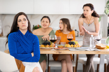Displeased miffed young brunette sitting arms folded at house get-together, with female friends in background rebuking her, expressing dissatisfaction and disapproval