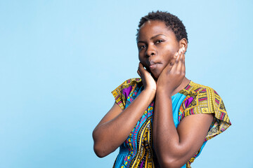 Fototapeta premium African american girl making silly facial expressions for the camera, feeling confident and carefree over blue background. Young joyful woman showing funny playful emotions, charming model.