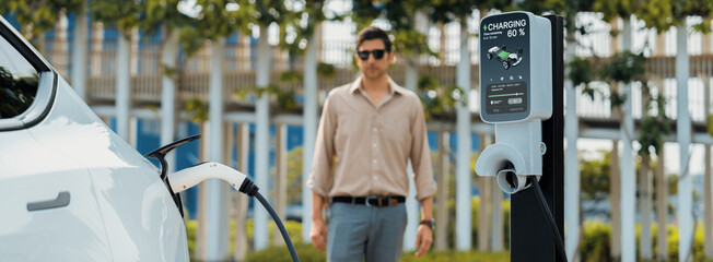 Young man recharge electric car's battery from charging station in outdoor green city park....
