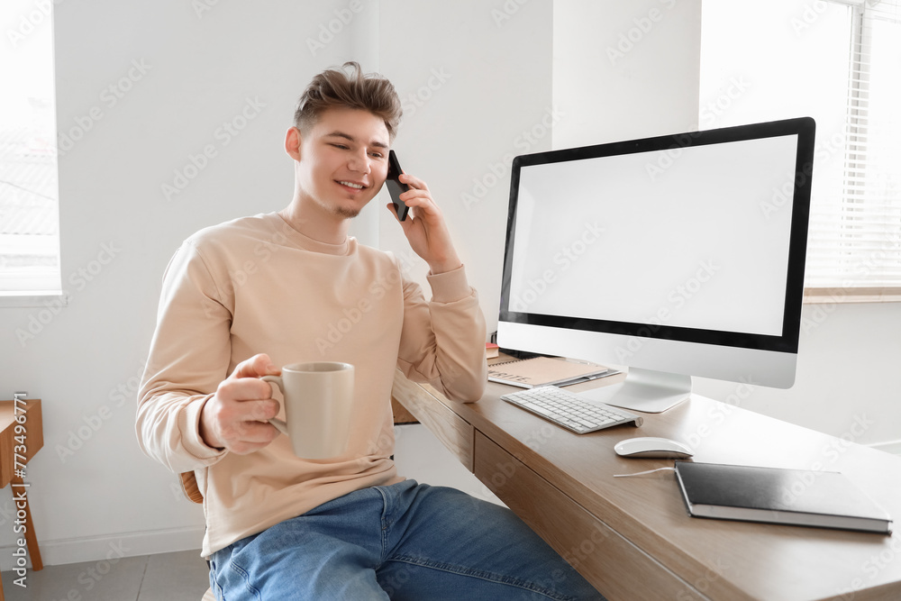 Poster young man with coffee cup talking by mobile phone at table in office