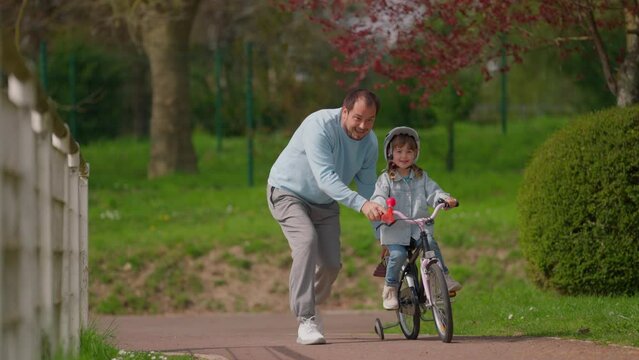 Little girl cycling in park with careful father who is teaching her to ride bicycle and is happy when she succeeds. Happy young family, fatherhood and childhood, active lifestyle concept