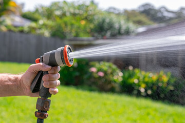 Garden hose with nozzle. Man's hand holding spray gun and watering plants, spraying water on grass in backyard.