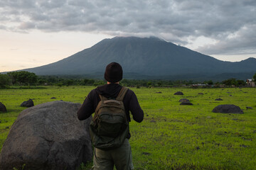 A male traveler in a knitted hat and backpack admires the view of the mountain, at sunrise, rear...