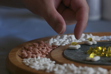 An addict's charcuterie board. Closeup of a hand selecting a white pill from a selection of prescription medications arranged on a cheese board as if they were an appetizer for those facing addiction.