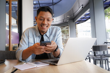 Asian man happy and smile face with blue shirt using laptop and mobile phone in coffee shop, online freelance business