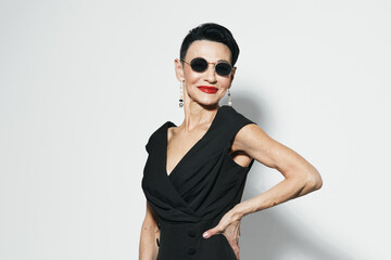 Fashionable woman in black dress and sunglasses posing in front of white wall for camera