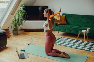 A woman is kneeling on a yoga mat in a plantfilled room, using an orange resistance band - obrazy, fototapety, plakaty