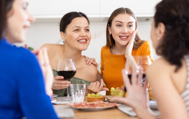 Four women friends are sitting at table, enjoying food and wine, chatting, talking, laughing. In...