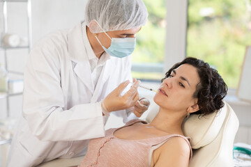 Middle-aged female patient lying on clinical chair undergoing face care procedure by injection method