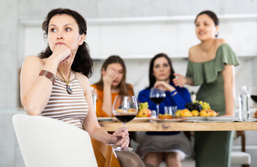 Displeased miffed woman sitting arms folded at house get-together, with female friends in background rebuking her, expressing dissatisfaction and disapproval - obrazy, fototapety, plakaty