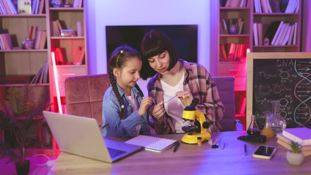 Mother with daughter looking at slides with plant cell in electron microscope at home. Focused young woman in casual wear sitting with little girl at table and using microscope for studying sciences.