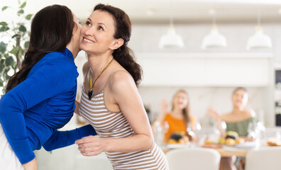 Relaxed and joyful dinner party scene at home, with smiling woman hosting friends, giving greeting kiss on cheek to bestie..
